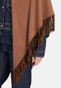 Poncho brun à franges porté sur une veste en denim bleu foncé et un jean, avec un accent sur le bas du torse et la main droite.