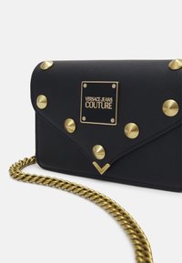 Clutch de cuero negro con tachuelas doradas y una correa de cadena dorada. Presenta una placa de logo cuadrada y un acento triangular en la parte inferior.