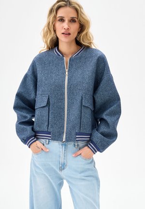 Kobieta o rozpuszczonych blond włosach nosi niebieską kurtkę na zamek z kieszeniami i prążkowanymi mankietami w paski, do tego jasnoniebieskie dżinsy, ręce w kieszeniach.