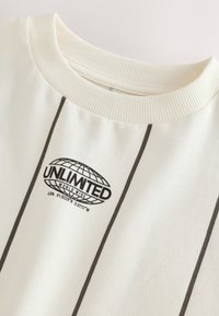Camisa blanca con rayas verticales negras, cuello redondo acanalado y un logo negro de un globo con el texto "UNLIMITED WORLD WIDE" y coordenadas debajo.