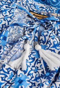Niebieska i biała tkanina w kwiatowy wzór, z zapięciem na wiązanie i białymi frędzlami z metalowymi akcentami. Widoczna metka marki.