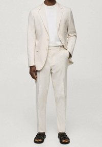 Svetle béžový oblek s texturovanou tkaninou, který zahrnuje blejzr s jedním knoflíkem, odpovídající kalhoty a bílé tričko. Černé sandály dokončují vzhled.