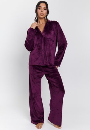 Mujer de pie descalza, vistiendo una camisa de botones de manga larga de terciopelo púrpura y pantalones anchos a juego, frente a un fondo blanco liso.