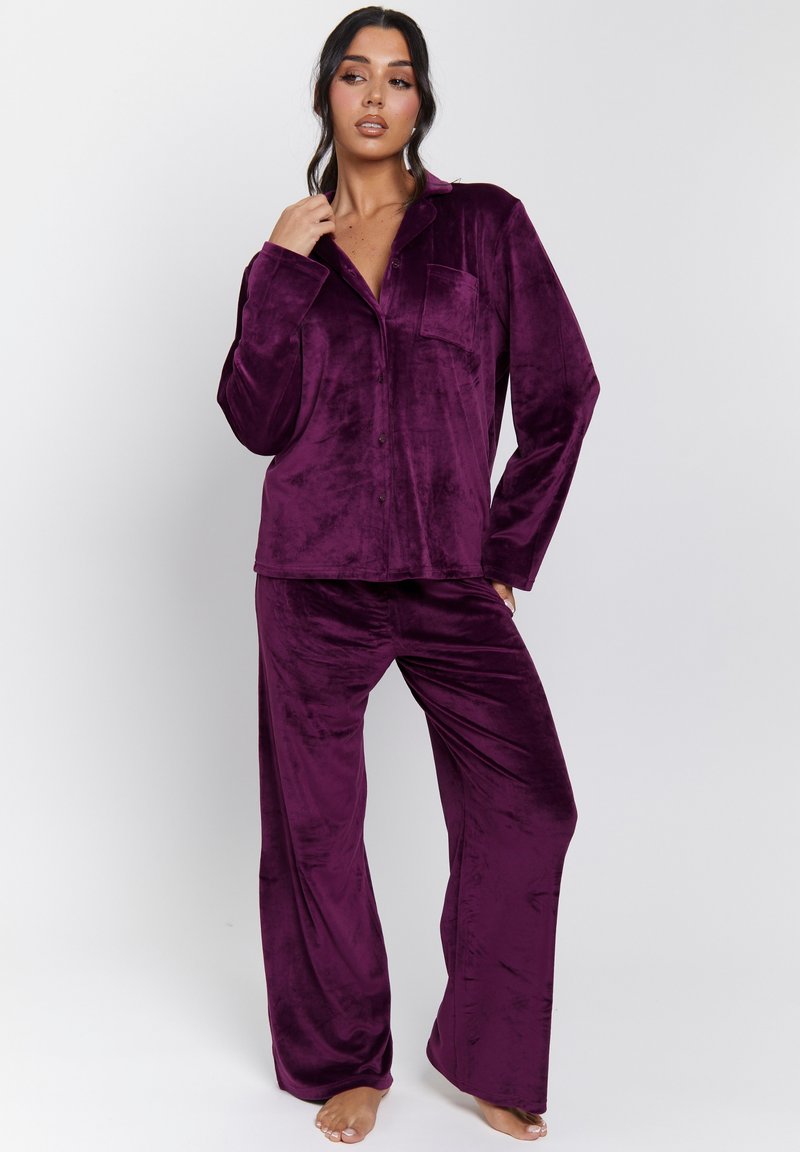 Kvinna står barfota klädd i en lila sammets långärmad skjorta med knappar och matchande vida byxor framför en plain vit bakgrund.