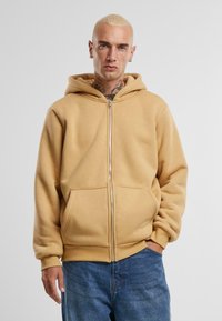 Bežová mikina na zips vyrobená z mäkkého materiálu, s prednou kapsou, voľným strihom a kapucňou. Pozadie je neutrálny šedý odtieň.