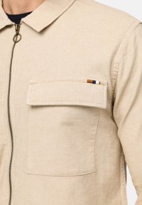 Veste beige en tissu texturé, dotée d'une fermeture éclair, d'un col et d'une poche poitrine unique avec des accents rayés.