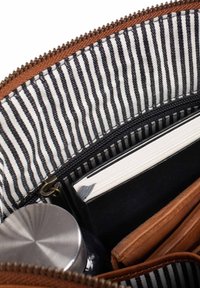 Innenraum eines braunen Täschchens mit Reißverschluss, ausgekleidet mit schwarz-weiß gestreiftem Stoff. Enthält eine Metallkappe, ein schwarzes Notizbuch und eine braune Geldbörse.