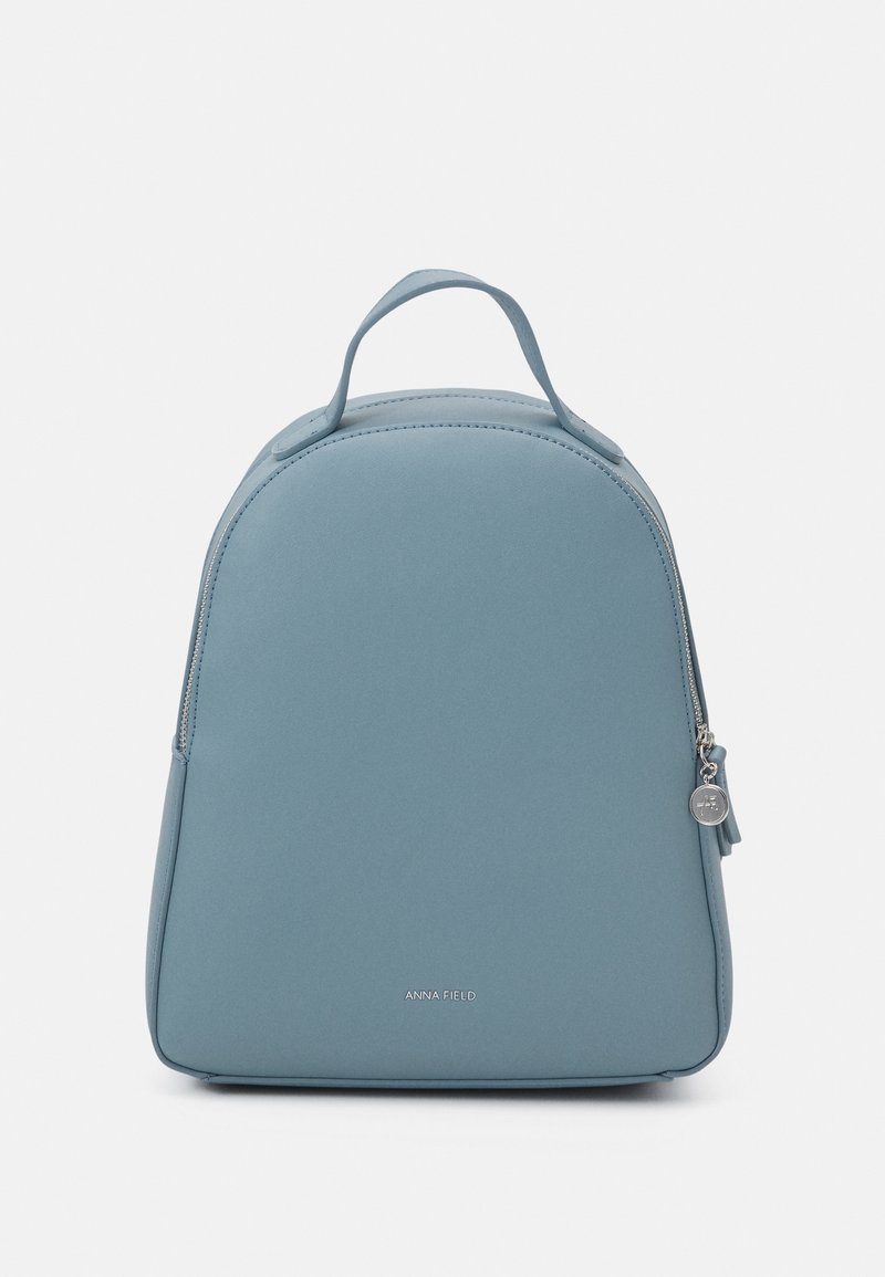 Petit sac à dos bleu clair avec un dessus arrondi, une poignée unique, une fermeture éclair et le logo "ANNA FIELD" à l'avant.