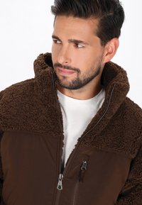 Mann mit dunklem Haar und Bart, der eine braune Fleecejacke mit hohem Kragen über einem weißen Hemd trägt und nach links schaut.