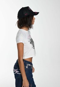 White cropped t-shirt with graphic print, paired with high-waisted blue jeans featuring pink lettering and a patch on the back pocket. Black cap.