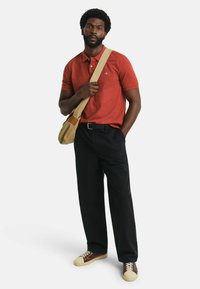 Rotes Polohemd, schwarze Hose, braune und cremefarbene Sneaker. Das Modell trägt eine hellbraune Umhängetasche mit einem Lederakzent.