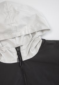 Czarna kurtka z białym kapturem, z zamkiem błyskawicznym z przodu i gładką fakturą. Projekt zawiera kontrastujące kolory i czyste linie.