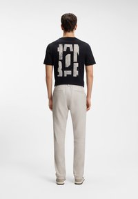 Hombre de espaldas, vistiendo una camiseta negra con texto en bloques blancos y pantalones beige claros, frente a un fondo blanco liso.