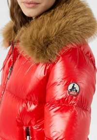 Giacca a gonfie rosso con un grande colletto di pelliccia marrone, soffice e voluminoso. Presenta una chiusura con zip e una toppa con il logo sulla manica. Texture liscia e lucida.