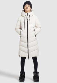 Lange witte pufferjas met een capuchon, voorzien van een ritssluiting aan de voorkant, zwarte accenten en chevronsteek. Draagt met zwarte leggings en laarzen.