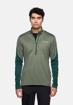 Giovane uomo che indossa una maglia sportiva verde a maniche lunghe con zip a quarto, con maniche di colore contrastante, in piedi davanti a uno sfondo chiaro e uniforme.