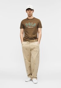 Άνδρας που φοράει καφέ μπλουζάκι Polo Ralph Lauren, μπεζ παντελόνι με μικρά κεντημένα σχέδια, λευκό καπέλο και λευκά αθλητικά παπούτσια, όρθιος με τα χέρια στις τσέπες.