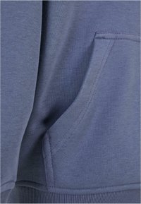 Felpa blu in tessuto con tasca frontale caratterizzata da dettagli di cucitura, orlo a coste e texture liscia. Vista ravvicinata del materiale e del design.