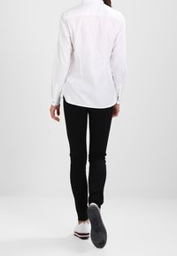Camisa blanca de botones con mangas largas, combinada con pantalones negros ajustados y zapatos blancos con un acento rojo y azul. Detalles de costura visibles.