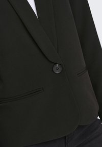 Blazer noir à un bouton avec revers à encoche et deux poches frontales horizontales, porté avec un pantalon foncé.