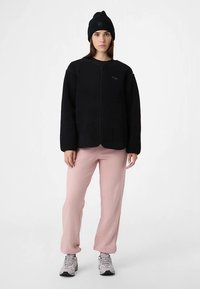 Zwarte fleecejas met ritssluiting aan de voorkant, lange mouwen en zakken, gecombineerd met losse, roze broek en een zwart gebreide muts.