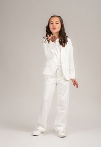 Junges Mädchen in einem weißen Anzug und Sneakers, das einen Kuss mit der Hand bläst, vor einem hellen, einfarbigen Hintergrund.