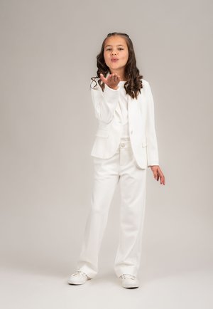 Junges Mädchen in einem weißen Anzug und Sneakers, das einen Kuss mit der Hand bläst, vor einem hellen, einfarbigen Hintergrund.