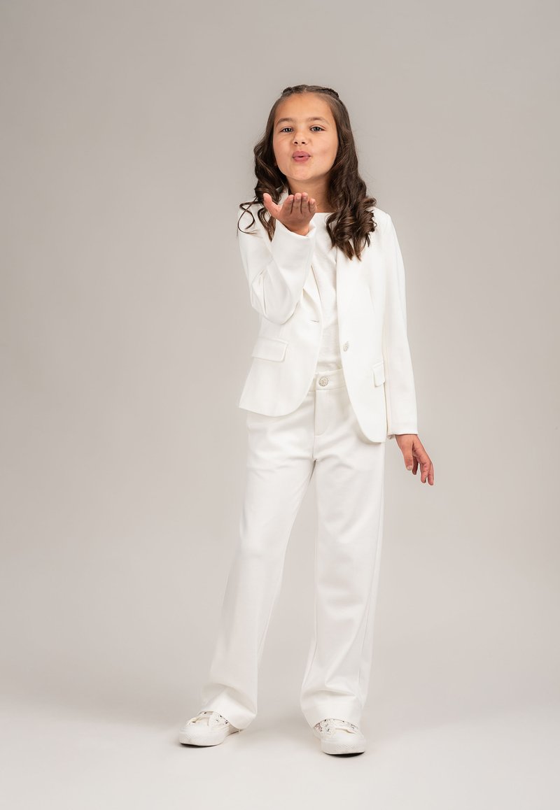 Junges Mädchen in einem weißen Anzug und Sneakers, das einen Kuss mit der Hand bläst, vor einem hellen, einfarbigen Hintergrund.