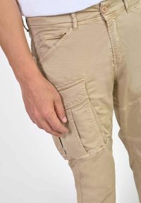 Lichtbeige cargobroek met meerdere zakken, waaronder een zijflapzak, gedragen door een persoon met een zichtbare linkerhand nabij de dij.