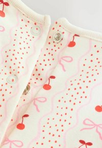 Close-up of cream-colored fabric with pink wavy lines, red dots, cherry prints, and metal snap buttons on a garment opening.