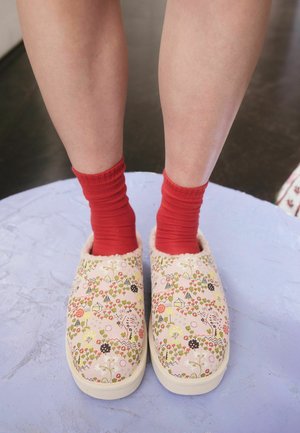 Benen gekleed in felrode sokken en slip-on pantoffels met bloemmotief, staand op een lichtpaarse gestructureerde ondergrond.