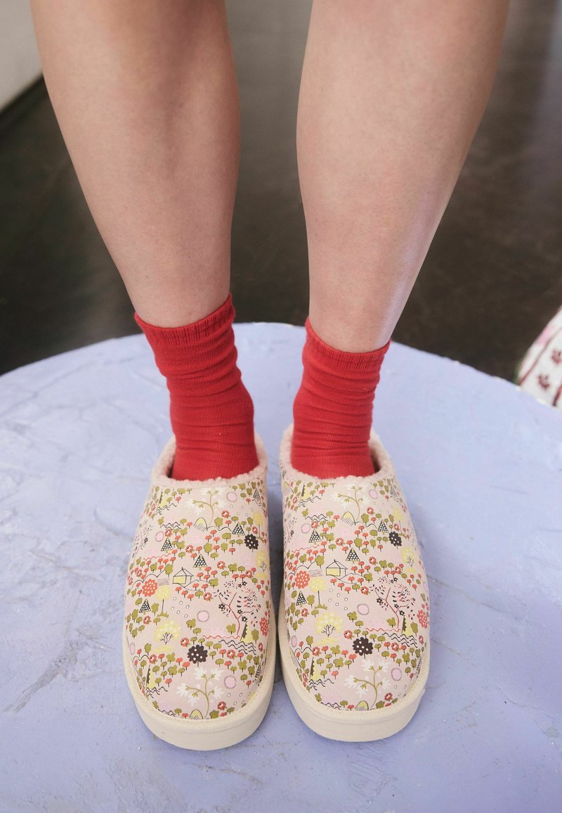 Benen gekleed in felrode sokken en slip-on pantoffels met bloemmotief, staand op een lichtpaarse gestructureerde ondergrond.