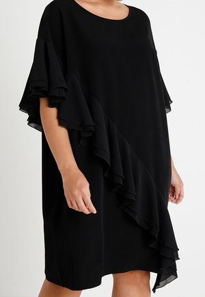 Løs sort kjole med lagdelte flæseærmer og diagonalt flæsessedetalje fra skulder til kant, båret af en person, der står.