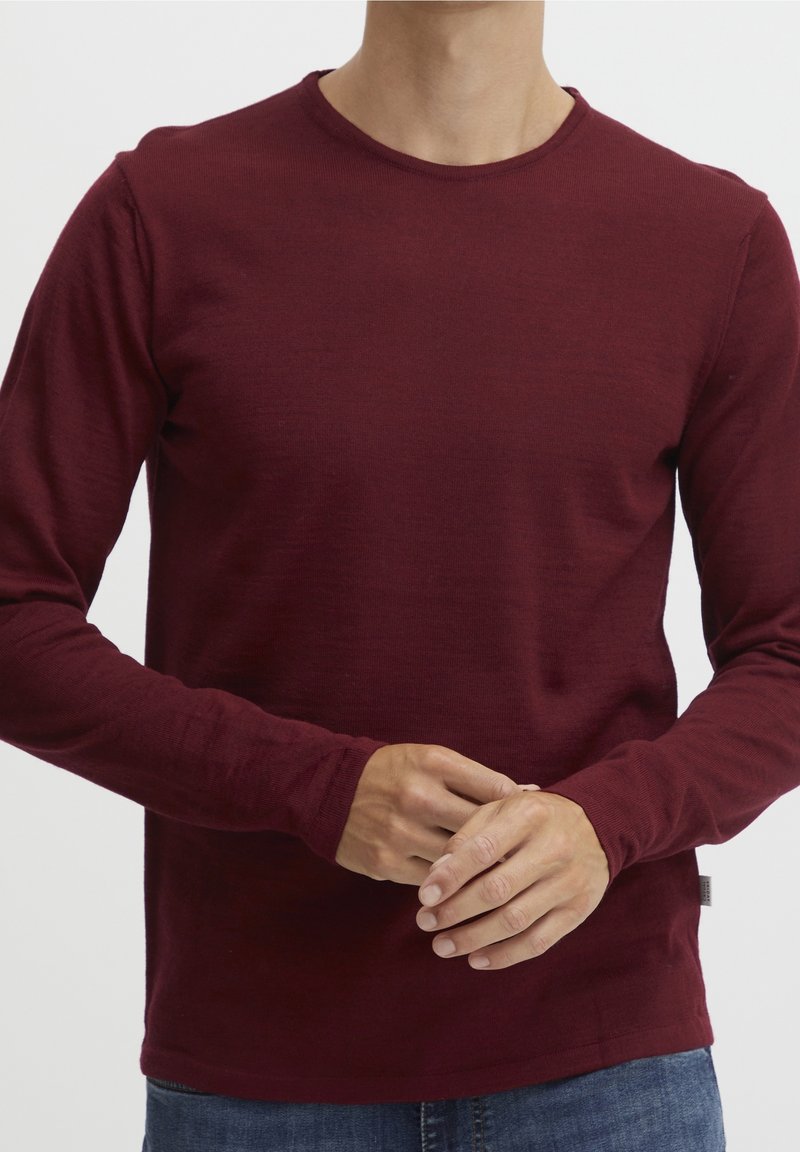 Homme portant un pull à col rond bordeaux à manches longues, les mains jointes devant, vu du cou aux hanches sur un fond blanc.