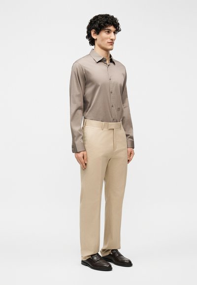 Νεαρός άνδρας που στέκεται φορώντας πουκάμισο ταμπά με κουμπιά, μπεζ παντελόνι με ζώνη και σκούρα καφέ δερμάτινα παπούτσια, σε απλό φωτεινό φόντο.