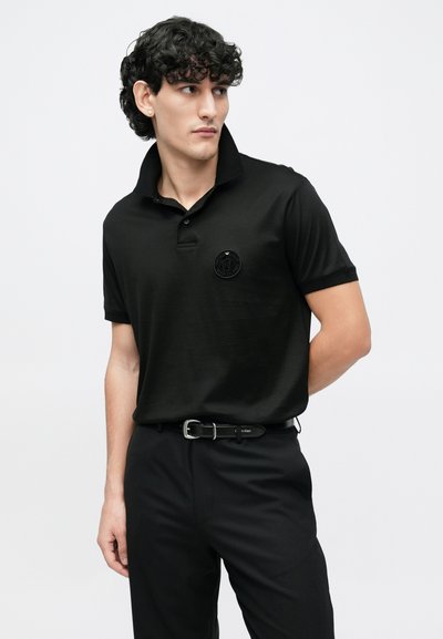 Juodas polo marškinėlis iš minkštos medžiagos, su smailėjančiu apykakliu ir juodu apskritu logotipu ant krūtinės. Dėvimas su juodomis kelnėmis ir diržu.