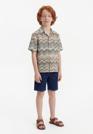 Red-haired boy stands wearing a multicolor zigzag patterned short-sleeve shirt, dark blue denim shorts, and brown leather sandals.