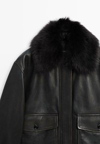 Giacca di pelle nera con un ampio colletto in pelliccia sintetica, dotata di due tasche frontali e una finitura testurizzata. Il design è elegante e moderno.