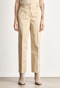 Άτομο που φοράει μπεζ παντελόνι ραμμένο στα μέτρα, μπεζ αμάνικη μπλούζα, μπεζ σουέντ μποτάκια μέχρι τον αστράγαλο, με τατουάζ λουλουδιών και αστεριών στο δεξί αντιβράχιο.