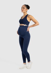 Soutien-gorge de sport de maternité bleu marine et leggings taille haute, avec une texture lisse et un design minimaliste. Porté avec des baskets blanches.