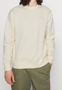 Sudadera amarilla clara hecha de un tejido suave, con cuello redondo, mangas largas, puños acanalados y un pequeño logo de Nike en color púrpura.