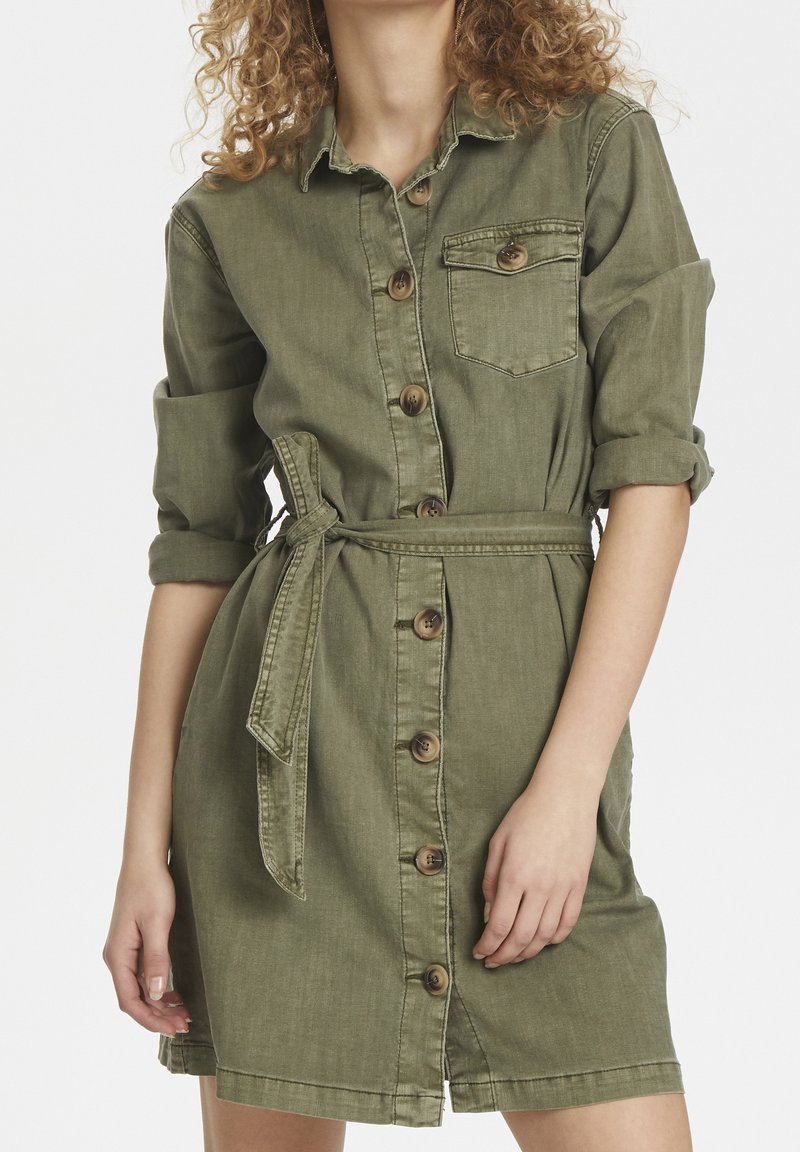 Robe en denim vert olive avec manches longues, bouton devant, une poche poitrine et une taille nouée, présentant une silhouette ajustée et des poignets retroussés.