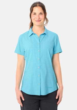 Frau trägt ein hellblaues, kurzärmeliges Hemd mit einem dezenten karierten Muster und schwarze Hose, steht vor einem einfarbigen Hintergrund.