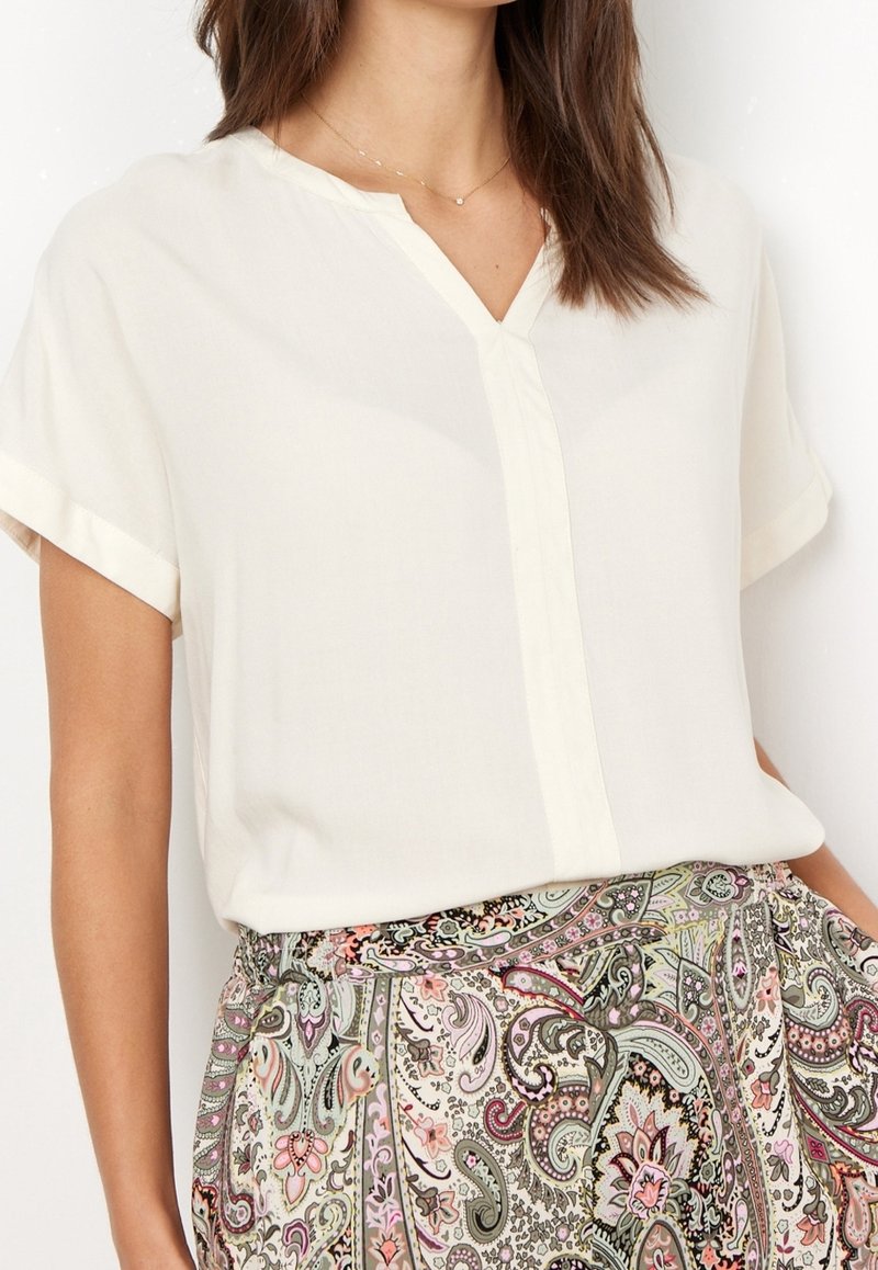 Blouse courte couleur crème à col en V avec des finitions contrastantes. Portée avec un pantalon taille haute à motif paisley multicolore.