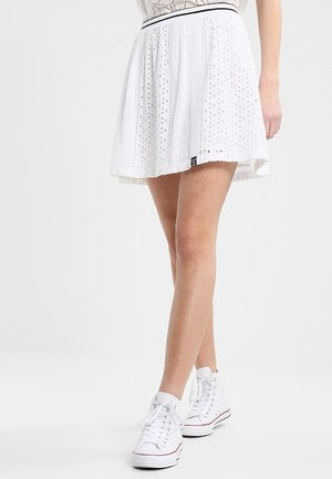 Jupe blanche avec des motifs découpés, design plissé et taille noire. Associée à des baskets montantes blanches.
