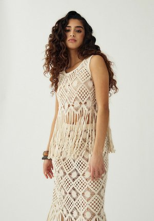 Femme aux longs cheveux bouclés portant un haut sans manches en crochet crème avec des franges et une jupe assortie, se tenant devant un fond uni.