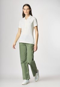 Maglietta polo bianca con un piccolo logo, abbinata a pantaloni larghi verde oliva e scarpe da ginnastica bianche. Design e colori semplici e casual.