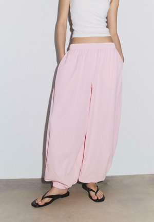 Osoba oblečená v voľných ružových nohaviciach s elastickými manžetami a pásom, v bielom tielku bez rukávov a čiernych žabkách stojaca pri jednofarebnej stene.