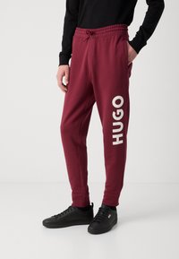 Bordeauxfarbene Jogginghosen mit einem Kordelzugbund, seitlichen Taschen und einem großen weißen "HUGO"-Logo am linken Bein; kombiniert mit schwarzen Sneakers.