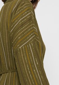 Veste olive à manches longues avec des rayures verticales en beige, marron et jaune. Texture douce, taille ceinturée et coupe décontractée.